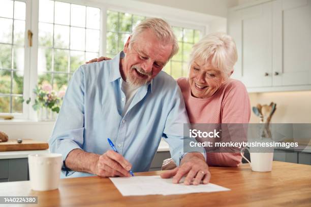 Retired Senior Couple Sitting In Kitchen At Home Signing Financial Document Stock Photo - Download Image Now