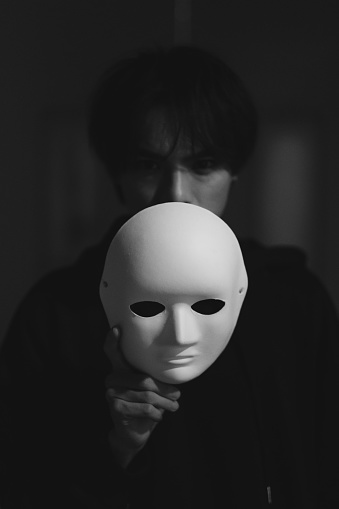A Halloween theme close-up portrait of a young man holding up a white empty mask with his hand covering his mouth or half of his face, as he looks at the camera.\n\nA moody horror portrait of a person with a confusing identity, expressing negative emotions of confusion, anxiety and fear.