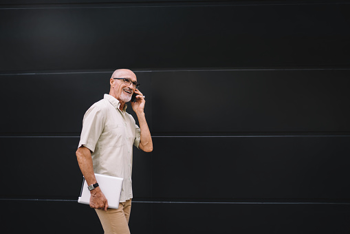 Positive bearded aged male with eyeglasses and tablet in hand talking on mobile phone while walking near modern black wall