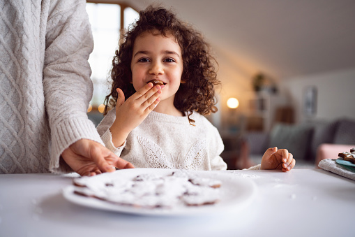 In the kitchen, Caucasian mother and daughter eating gingerbread cookies they made, while enjoying Christmas tradition