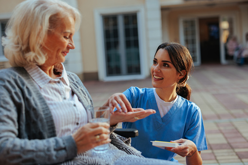 A young adult Caucasian caregiver is handing out medication to her elderly patient in the nursing home yard.