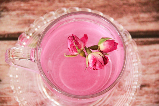 Tea made from pink rose petals in a transparent glass mug and small flower buds on  wooden background