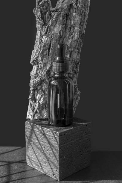 istock Glass bottle with dropper lid on square 3d platform leaning against piece of tree bark. Unmarked container with moisturizing oil on black background in beam of light. Vertical image, monochrome 1440244387