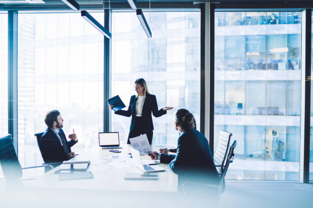 Confident female director dressed in formal suit making presentation of productive strategy near desktop with laptop computer with blank screen area for advertising text or financial website