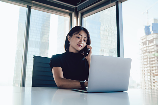 Concentrated Asian businesswoman using laptop and speaking on smartphone