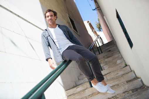 Portrait of young Caucasian man having fun in summer city. Happy man sitting on metal railing like trying to slide downstairs and looking aside. Active rest, vacation, leisure and travelling concept