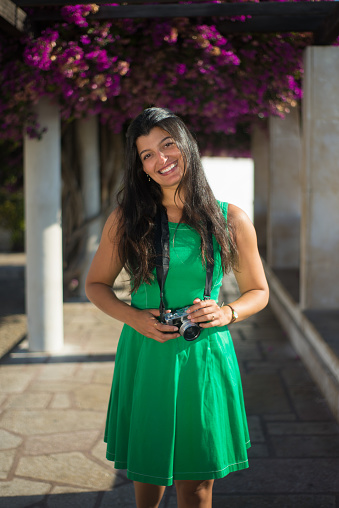 Portrait of beautiful woman with photo camera. Young woman travelling in old city standing on bougainvillea trees alley holding camera looking straight and smiling. Travelling and active rest concept