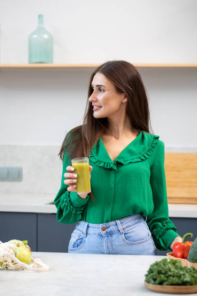 привлекательная брюнетка стоит на кухне со стаканом зеленого смузи. здоровое питание, вегетарианство. - apple women green eating стоковые фото и изображения
