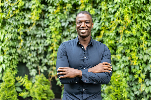 A confident African-American man standing in a garden against a leafy wall with his arms crossed and smiling at the camera
