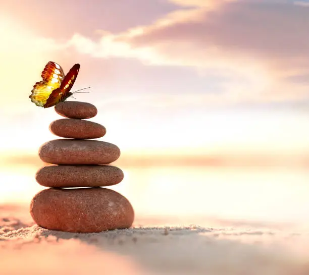 Photo of Balanced pebble pyramid and butterfly on the beach. Abstract warm sunset bokeh with sand on the background. Zen stones on the sea beach.