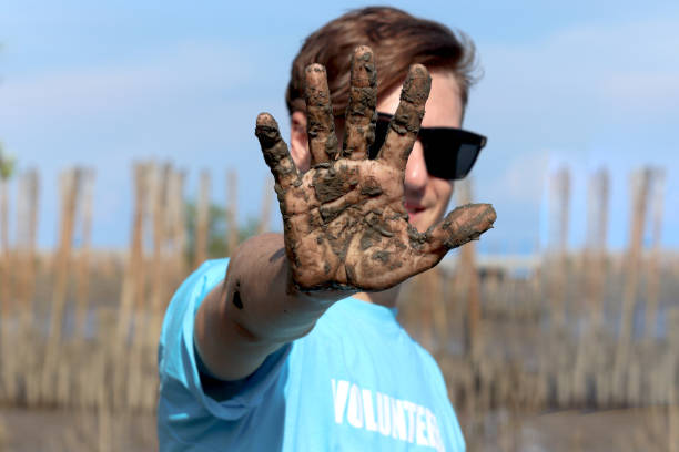 Volunteer young man in blue t-shirt showing hand covered mud dirty after planting sapling tree in deep mud at mangrove forest for increase mangrove cover world-wide, Eco world environment day concept. joven voluntario con camiseta azul que muestra barro cubierto a mano sucio después de plantar árboles jóvenes en lodo profundo en el bosque de manglares para aumentar la cobertura de manglares en todo el mundo, concepto del día mundial del medio ambien