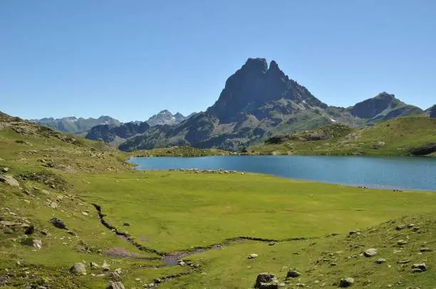 A low-angle of a landscape with Lacs d'Ayous lake view, meadow around, and ridged mountain peak against clear sky background