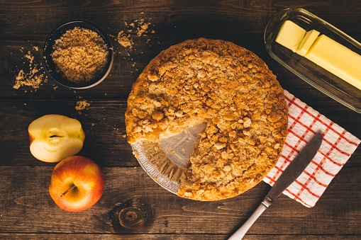 Fall baking scene with an apple pie with a piece missing. Scene includes butter, sliced apple, a bowl full of brown sugar and a butter knife.