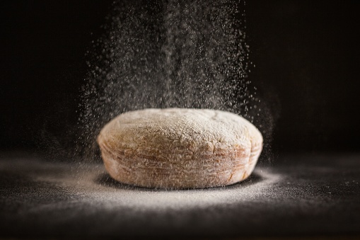 Flour being sprinkled on freshly baked bread with a dark background. Perfect for cooking-related articles or posts