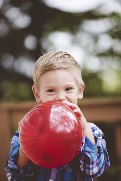 toma vertical de un niño feliz inflando un globo rojo con un fondo borroso - balloon blowing inflating child fotografías e imágenes de stock