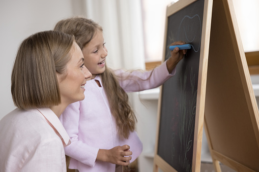 Young woman and daughter drawing on blackboard use chalk, enjoy creative hobby together. Art class for preschool children, daycare, caring parent teach kid, share skills spend leisure at home concept