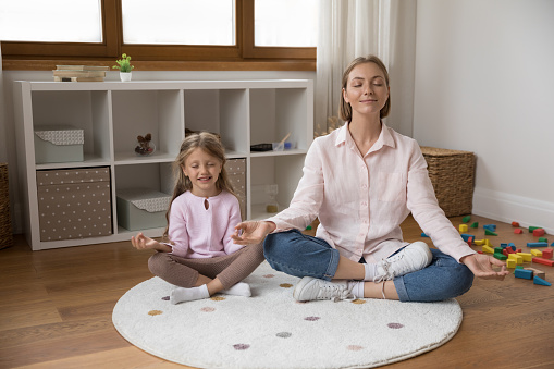 Beautiful young mom meditating with little peaceful daughter, sitting cross-legged on carpet in cozy playroom. Loving parent teach child healthy life habit, doing yoga practice at home, rest concept