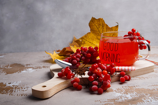A glass cup with viburnum fruit tea and ripe red viburnum berries on gray background, autumn morning tea concept, good morning, selective focus, copy space.