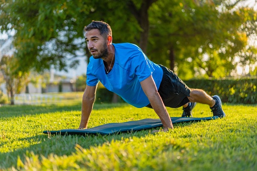 Hispanic man in blue t-shirt and mat doing push-ups in the park garden during sunset light.