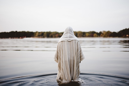 A person wearing a biblical robe walking in the water with a blurred background shot from behind