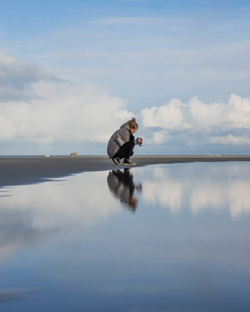 vista vertical de una mujer tomando una foto en la playa con su reflejo en el agua - mujer agachada perfil fotografías e imágenes de stock