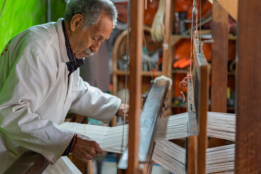 An older man, an artisan by profession, is in his factory where he weaves handcrafted looms.