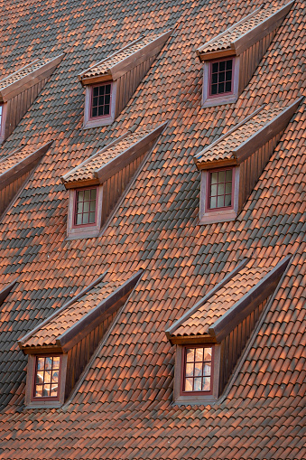 Vertical shot of closed attic or mansard windows on the rooftop covered with ceramic tiles. House with brown clay shingles and skylights on roof. Building construction concepts