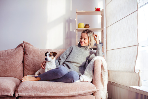 Beautiful middle-aged woman with dog at home sitting on couch in front of window, relaxing in her living room