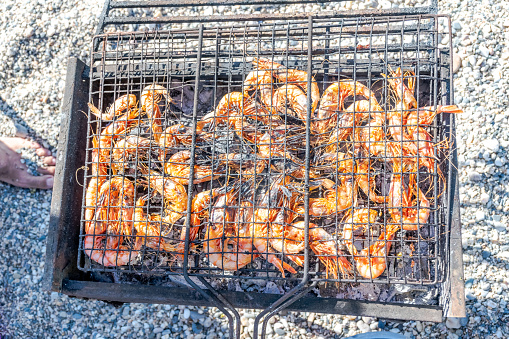 Overhead view of grilled shrimp in a barbecue grill, a foot with blurred beach pebbles  on background.