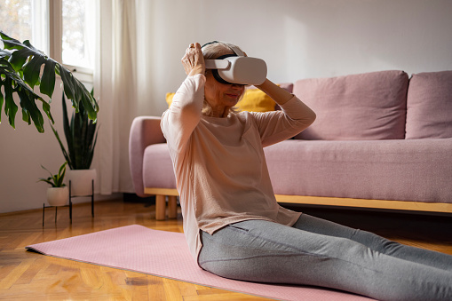Senior woman in the morning doing yoga with virtual reality headset in a cosy living room