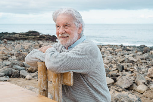 Smiling relaxed mature senior man with beard sitting in outdoors at sea. White haired caucasian elderly grandfather sitting at the beach on a wooden chair with crossed arms
