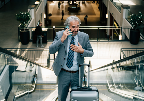 Happy business man in elegant gray suit speaking on his phone while moving up escalator with his luggage at the airport.
