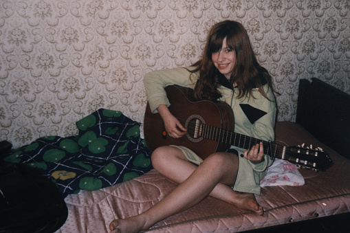 Teenage girl is sitting on bed and playing guitar. Vintage home photo