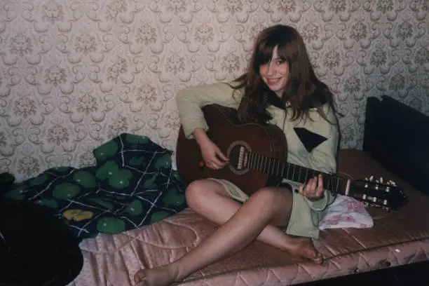 Teenage girl is sitting on bed and playing guitar. Vintage home photo