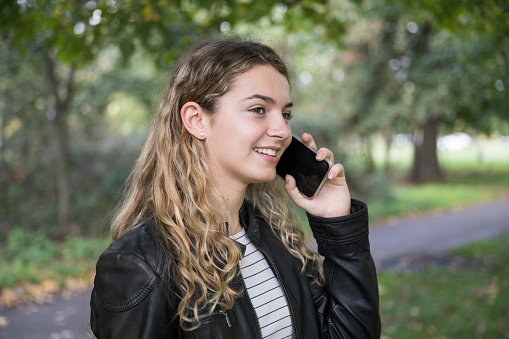 Three-quarter front view of young Caucasian woman with long blond hair, casually dressed for autumn weather, and smiling while communicating with caller.