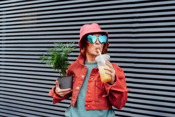 mujeres de moda hipster con ropa brillante, gafas en forma de corazón, sombrero de cubo bebiendo té de burbujas de perla de tapioca con sabor a azúcar afrutado y sosteniendo una planta verde en maceta sobre el fondo de la pared a rayas grises - people clothing elegance built structure fotografías e imágenes de stock