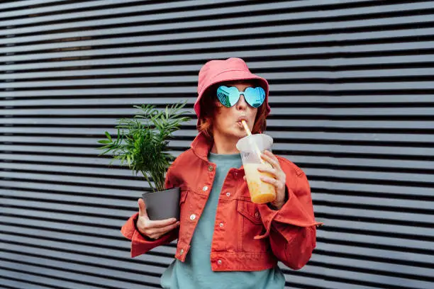 Hipster fashion women in bright clothes, heart shaped glasses, bucket hat drinking fruity sugar flavored tapioca pearl bubble tea and holding green potted plant on the gray striped wall background.