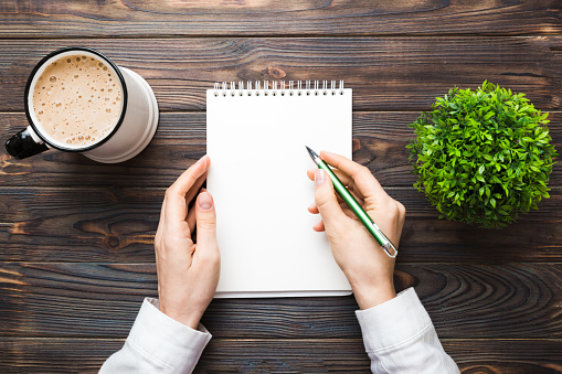 Woman hand with pencil writing on notebook and hold coffee cup. Woman working on office table with coffee.