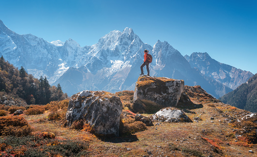 Standing woman with backpack on the stone and beautiful mountains at sunset. Landscape with sporty girl, high rocks, snowy peaks, blue sky in autumn. Travel in Nepal. Lifestyle. Trekking in Himalayas