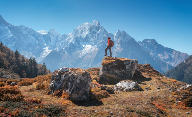 Standing woman with backpack on the stone and beautiful mountains at sunset. Landscape with sporty girl, high rocks, snowy peaks, blue sky in autumn. Travel in Nepal. Lifestyle. Trekking in Himalayas stock photo