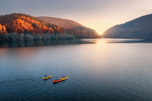 Aerial view of people on floating colorful boats on blue lake in mountains with red forest at sunset in autumn. River in carpathian mountains in fall in Ukraine. Landscape. Top view of canoe. Travel
