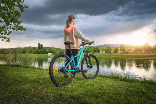 Woman riding a mountain bike near lake at overcast sunset in spring. Colorful landscape with sporty girl, bicycle, coast of river, green grass, cloudy sky in park in summer. Sport and travel. Biking