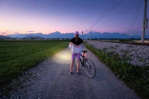 Woman is standing with mountain bike on gravel road at night in summer. Colorful landscape with sporty girl, bicycle with light, field, dirt road, green grass, purple sky at sunset. Sport and travel