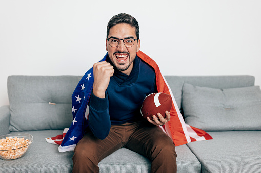 Excited fan watching rugby game on TV while sitting on sofa with sports ball and American flag