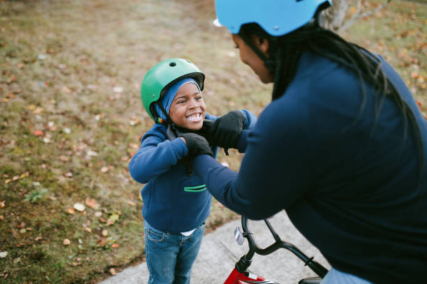 Mother And Son Preparing for Bike Ride A young African American mom helps her son strap his bike helmet on, a fun time of family exercise, riding bikes together on a cool Autumn day. Healthy lifestyle and good childhood memories. cycling helmet stock pictures, royalty-free photos & images