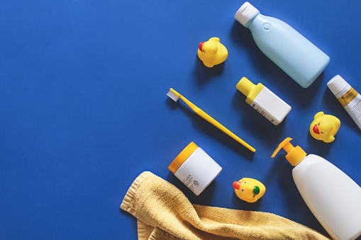 Baby shower. Baby bathing accessories on blue table. White and yellow shampoo bottle, yellow towel, comb, toothbrush, baby cream and yellow rubber ducks. Flat lay. Top view. Copy space