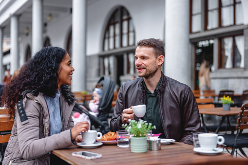 Waist-up side shot with blurred background of a mid-adult Caucasian man talking to his girlfriend about future plans together and holding a cup of coffee while she listens attentive to him, both seated in an outdoors cafeteria enjoying breakfast.