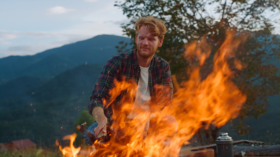 Camping hipster watch fire in nature forest. Happy guy sit bonfire in mountains close up. Wanderlust tourist traveling on beautiful landscape. Positive traveler enjoy evening. Activity leisure concept