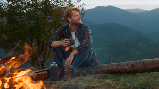 Travel man rest campfire on nature. Close up carefree guy enjoy mountains evening outside. Red hair man drink tea on vacation tourism activity. Relaxed hiker camping in forest. Tourist holiday concept