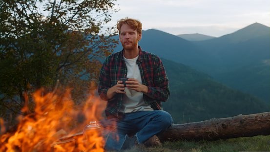 Camping hipster enjoy drink on mountains holiday. Young traveler relax on nature campfire close up. Ginger guy spend vacation outdoors. Chill tourist look burning fire on evening. Weekend concept.
